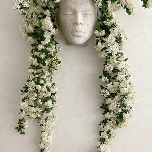 Pu&ograve; includere: Un vaso da fiori in ceramica bianca a forma di viso con un'espressione serena, circondato da una cascata di delicati fiori bianchi e foglie verdi.