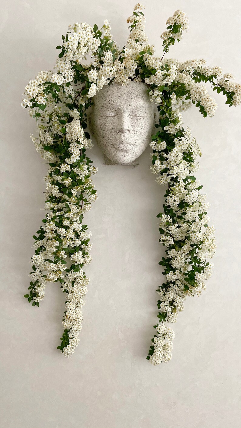 Pu&ograve; includere: Un vaso da fiori in ceramica bianca a forma di viso con un'espressione serena, circondato da una cascata di delicati fiori bianchi e foglie verdi.