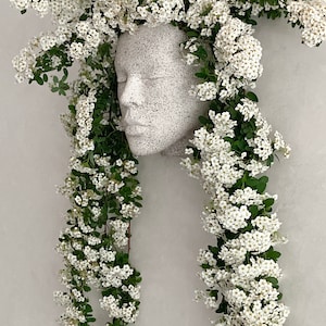 Pu&ograve; includere: Un busto in gesso bianco di un viso con fiori bianchi a cascata sui lati, creando un effetto di corona floreale.
