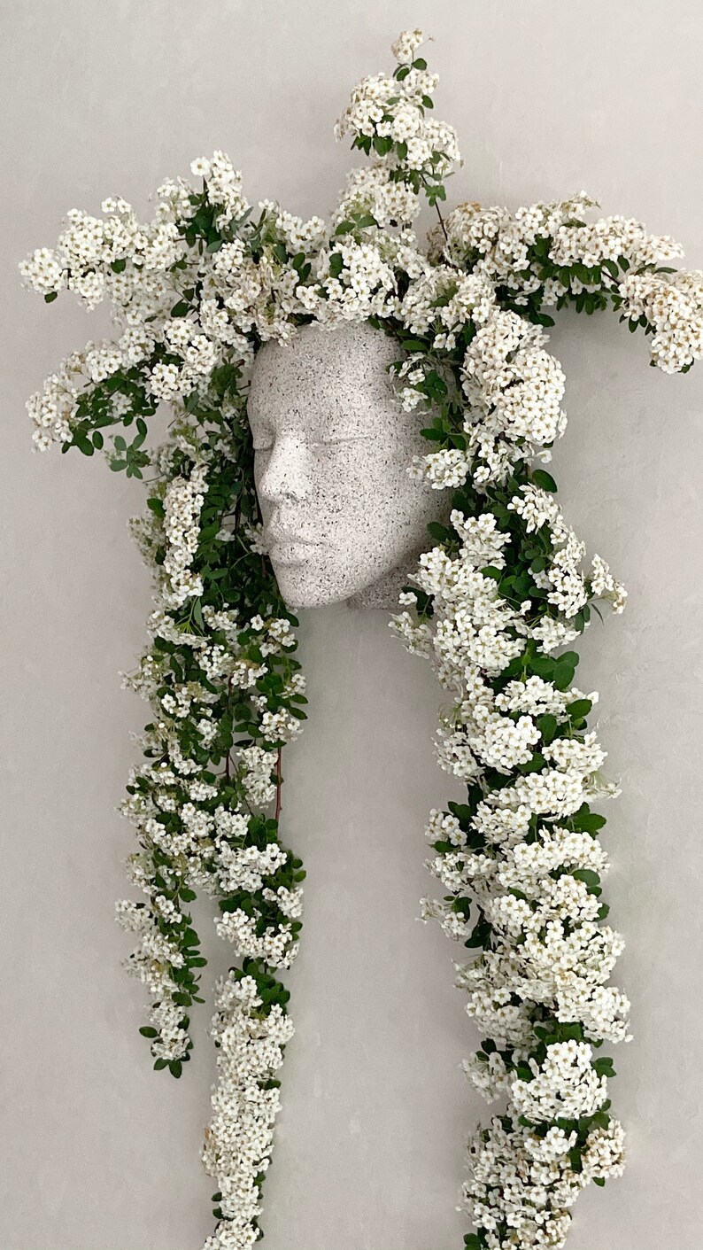 Pu&ograve; includere: Un busto in gesso bianco di un viso con fiori bianchi a cascata sui lati, creando un effetto di corona floreale.