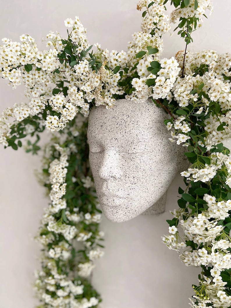 Pu&ograve; includere: Un vaso da fiori in ceramica bianca a forma di viso umano con fiori bianchi a cascata intorno.