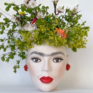 Puede incluir: Un macetero de cerámica blanca con forma de rostro de mujer con una corona de hojas verdes y flores blancas. El macetero tiene pendientes de flores rojas y una boca roja pintada.
