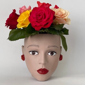 Könnte beinhalten: Ein Keramik-Pflanztopf in Form eines Frauenkopfes mit roten Rosen, gelben Rosen und cremefarbenen Rosen im Haar. Der Topf hat rote Ohrringe.