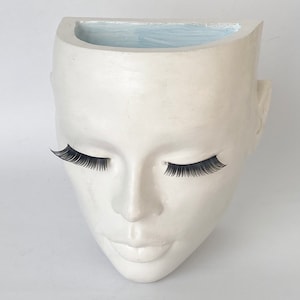 Puede incluir: Maceta de cerámica blanca con forma de cara con los ojos cerrados y pestañas largas. La maceta tiene un interior azul claro.