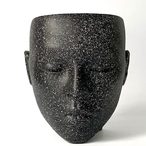 May include: A black ceramic planter shaped like a human face with closed eyes. The planter has a speckled texture.