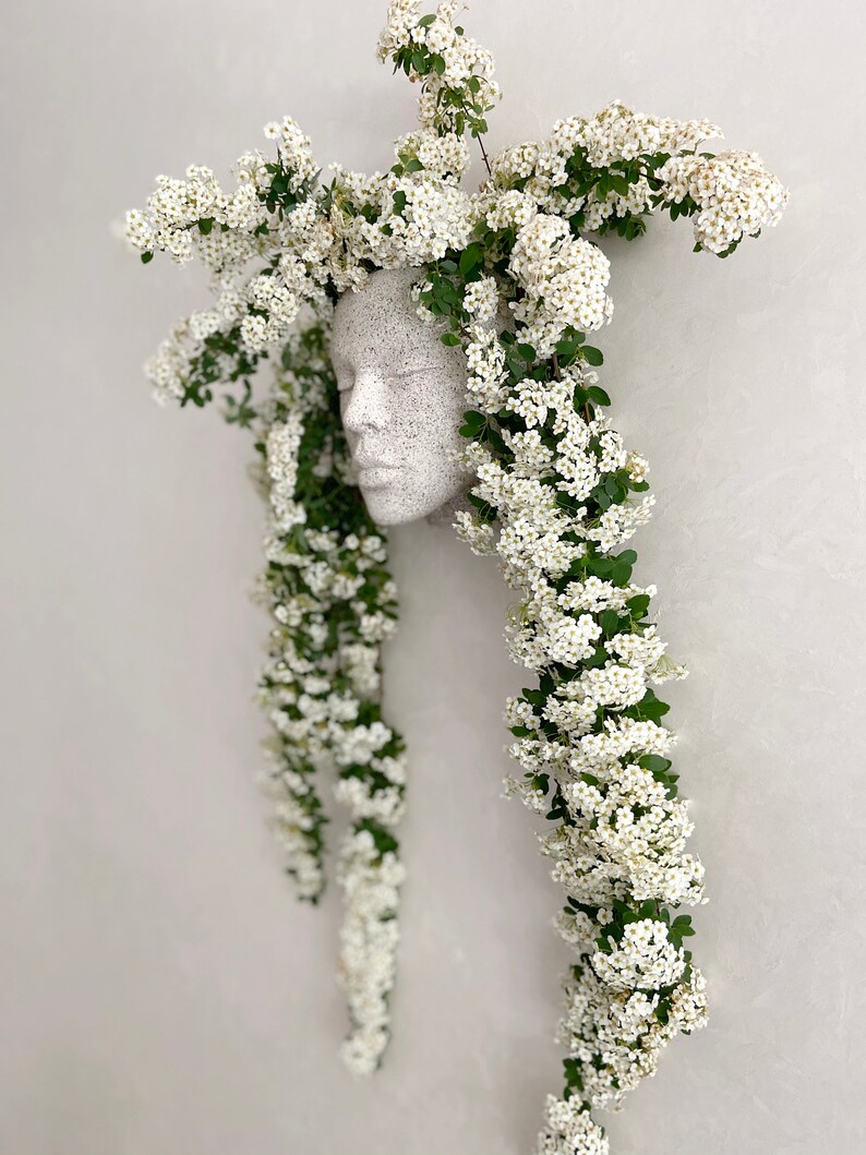 Pu&ograve; includere: Una scultura in gesso bianco di un viso con fiori bianchi a cascata dalla cima della testa.