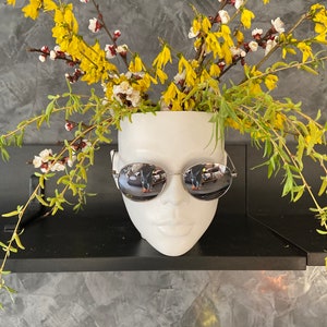 Pu&ograve; includere: Un vaso di ceramica bianca a forma di viso con occhiali da sole, pieno di fiori gialli.