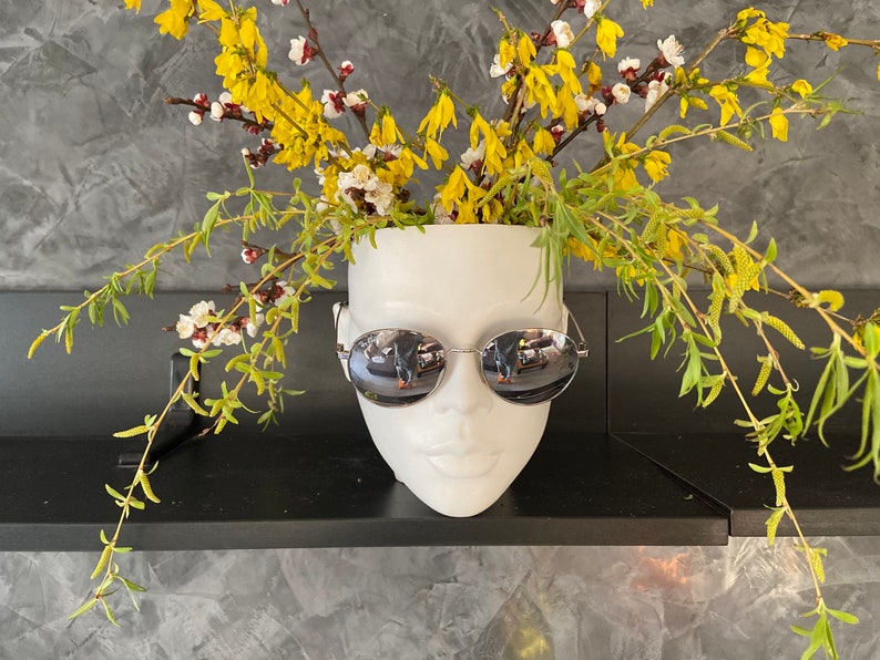 Pu&ograve; includere: Un vaso di ceramica bianca a forma di viso con occhiali da sole, pieno di fiori gialli.