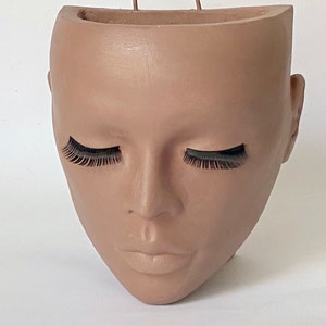 May include: A beige plaster planter shaped like a human face with closed eyes and eyelashes. The planter has a small opening at the top for planting.