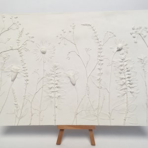 May include: White plaster relief art featuring a variety of pressed flowers and plants, displayed on a small wooden easel.