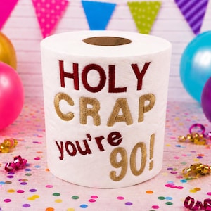 May include: A white roll of toilet paper featuring embroidered text in dark red and gold thread. The text reads HOLY CRAP you're 90! in a mix of block and script lettering. The roll is centred and stands upright against a festive party background.