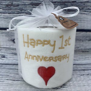 May include: A white toilet paper roll with gold embroidered text reading "Happy 1st Anniversary" and a red heart. The roll is wrapped in a white tulle bag and tied with a bow. A small tag is attached.