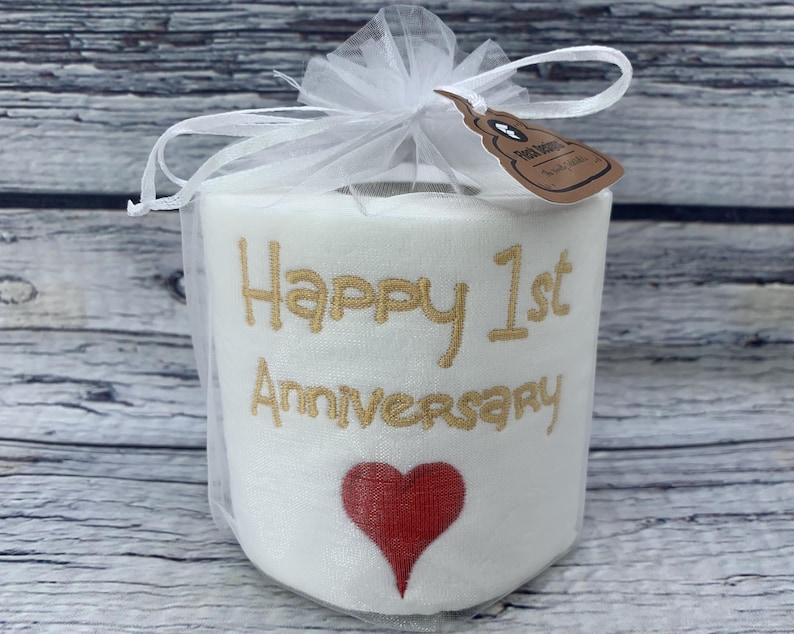 May include: A white toilet paper roll with gold embroidered text reading "Happy 1st Anniversary" and a red heart. The roll is wrapped in a white tulle bag and tied with a bow. A small tag is attached.