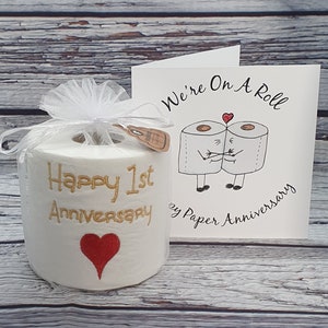 May include: A novelty gift set for a 1st anniversary. It includes a roll of toilet paper with "Happy 1st Anniversary" embroidered in gold, a red heart, and a card with a cartoon of two toilet paper rolls hugging, with the text "We're On A Roll".