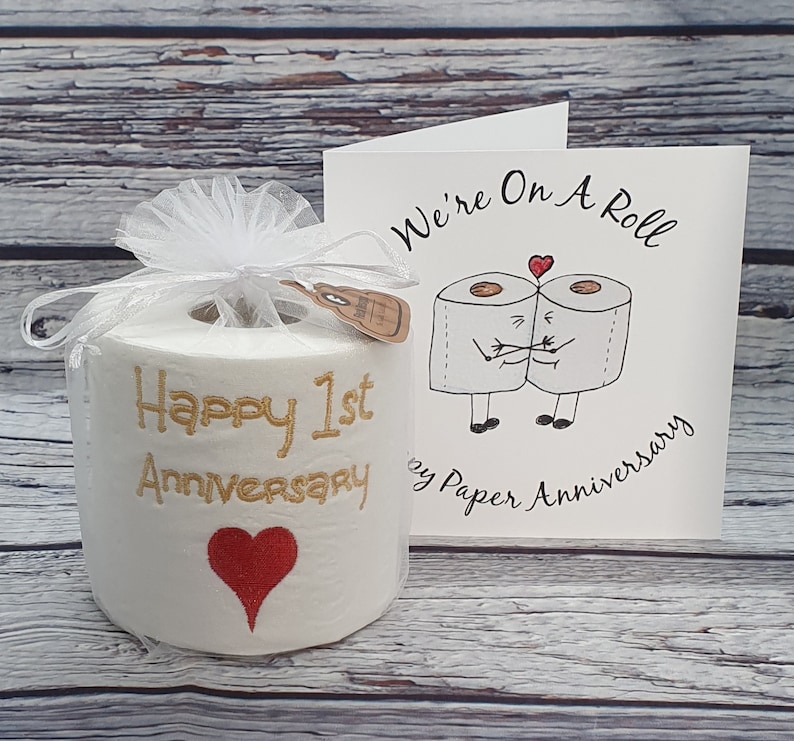 May include: A novelty gift set for a 1st anniversary. It includes a roll of toilet paper with "Happy 1st Anniversary" embroidered in gold, a red heart, and a card with a cartoon of two toilet paper rolls hugging, with the text "We're On A Roll".