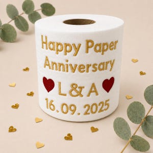 May include: A white toilet paper roll featuring gold embroidered text that reads Happy Paper Anniversary, the initials L & A, and the date 16.09.2025. Two red embroidered hearts are positioned on either side of the initials on the front surface.