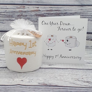 May include: A white toilet paper roll with "Happy 1st Anniversary" in gold script and a red heart. A card with cartoon toilet paper characters and the text "One Year Down... Forever to go!" is next to it. The roll is wrapped in a white mesh.