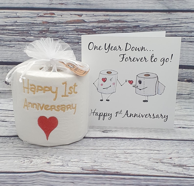 May include: A white toilet paper roll with "Happy 1st Anniversary" in gold script and a red heart. A card with cartoon toilet paper characters and the text "One Year Down... Forever to go!" is next to it. The roll is wrapped in a white mesh.