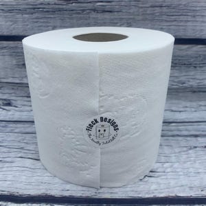 May include: A white roll of toilet paper with a black and white sticker that says "Fleck Designs. The smelly toilet co."