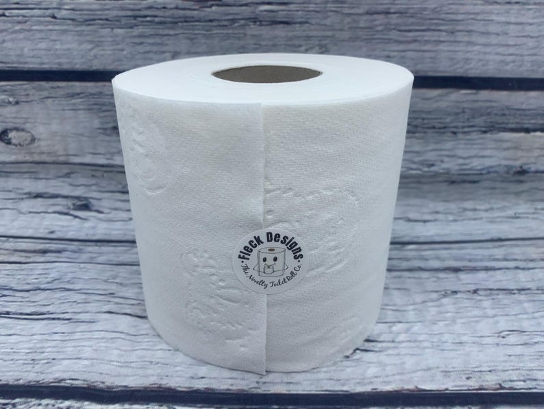 May include: A white roll of toilet paper with a black and white sticker that says "Fleck Designs. The smelly toilet co."