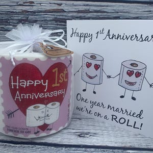 May include: A pink and white toilet paper roll with a heart and the text "Happy 1st Anniversary" and "One Year Down...Forever To Go!"  A white card with the text "Happy 1st Anniversary" and "One year married we're on a ROLL!"  The card features a cartoon image of two toilet paper rolls with faces and hearts for eyes.