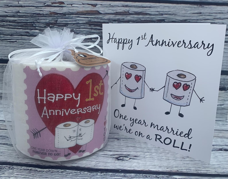 May include: A pink and white toilet paper roll with a heart and the text "Happy 1st Anniversary" and "One Year Down...Forever To Go!"  A white card with the text "Happy 1st Anniversary" and "One year married we're on a ROLL!"  The card features a cartoon image of two toilet paper rolls with faces and hearts for eyes.