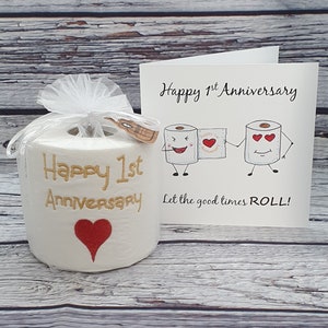 May include: A novelty gift set for a first anniversary. It includes a roll of toilet paper with "Happy 1st Anniversary" embroidered in gold, a red heart, and a greeting card with cartoon toilet paper characters.