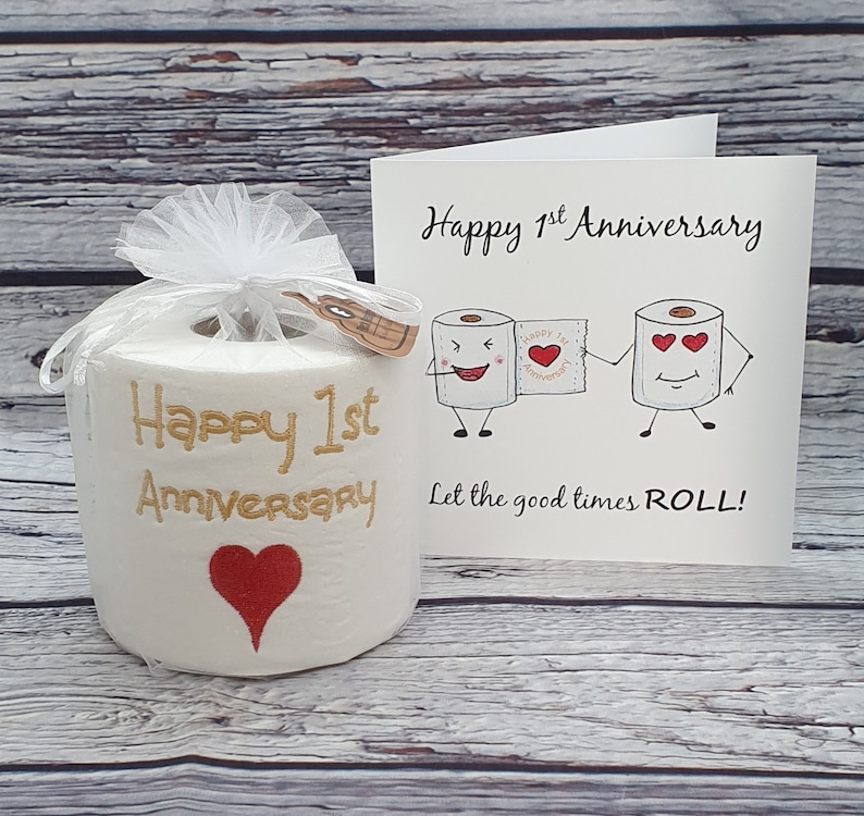 May include: A novelty gift set for a first anniversary. It includes a roll of toilet paper with "Happy 1st Anniversary" embroidered in gold, a red heart, and a greeting card with cartoon toilet paper characters.