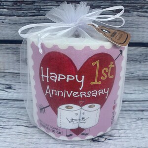 May include: A pink and white toilet paper roll with a red heart and the text "Happy 1st Anniversary" on the front. Two cartoon toilet paper rolls are holding hands below the text. The text "One year down...forever to go!" is printed below the cartoon rolls.