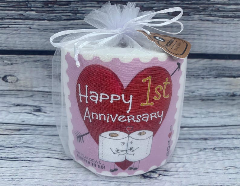 May include: A pink and white toilet paper roll with a red heart and the text "Happy 1st Anniversary" on the front. Two cartoon toilet paper rolls are holding hands below the text. The text "One year down...forever to go!" is printed below the cartoon rolls.
