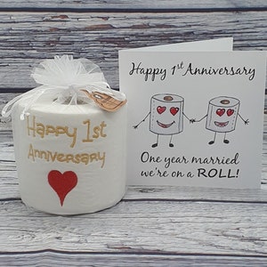 May include: A white toilet paper roll with "Happy 1st Anniversary" in gold script and a red heart. It is wrapped in a white mesh and tied with a ribbon. A card with cartoon toilet paper rolls holding hands reads "One year married we're on a ROLL!"