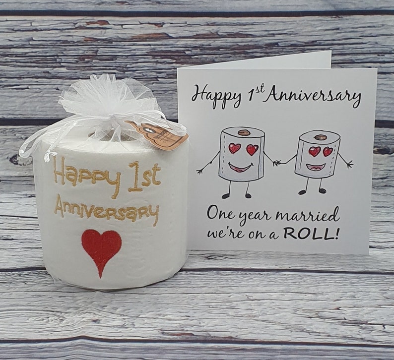 May include: A white toilet paper roll with "Happy 1st Anniversary" in gold script and a red heart. It is wrapped in a white mesh and tied with a ribbon. A card with cartoon toilet paper rolls holding hands reads "One year married we're on a ROLL!"