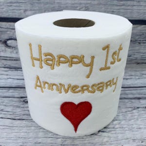 May include: White toilet paper roll with gold embroidered text reading "Happy 1st Anniversary" and a red heart. The roll is displayed against a light wood grain background. A unique gift for a first anniversary.