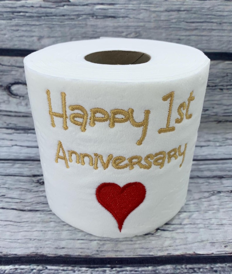 May include: White toilet paper roll with gold embroidered text reading "Happy 1st Anniversary" and a red heart. The roll is displayed against a light wood grain background. A unique gift for a first anniversary.