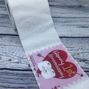 May include: A pink and red card with a heart and the text "Happy 1st Anniversary I love you" and two toilet paper rolls with faces on them. The card is attached to a roll of toilet paper.