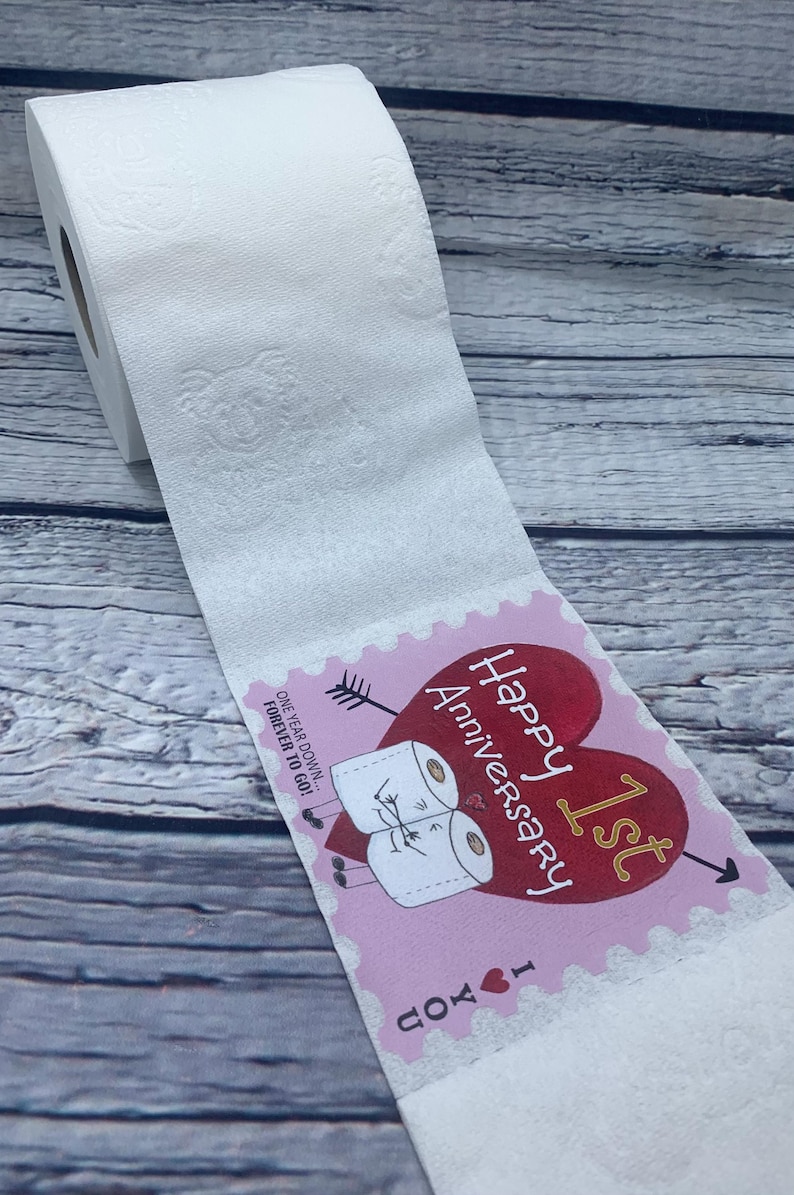 May include: A pink and red card with a heart and the text "Happy 1st Anniversary I love you" and two toilet paper rolls with faces on them. The card is attached to a roll of toilet paper.
