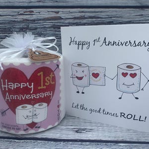 May include: A pink and red toilet paper roll with the text "Happy 1st Anniversary" and a heart. The roll is wrapped in a clear plastic bag with a white ribbon. A card with the text "Happy 1st Anniversary" and a cartoon of two toilet paper rolls holding hands is in the background.