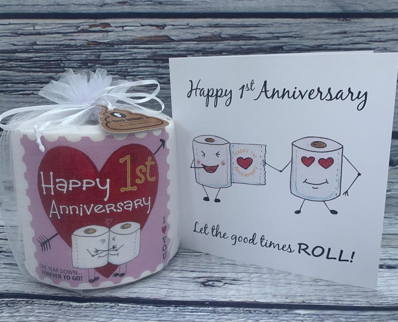 May include: A pink and red toilet paper roll with the text "Happy 1st Anniversary" and a heart. The roll is wrapped in a clear plastic bag with a white ribbon. A card with the text "Happy 1st Anniversary" and a cartoon of two toilet paper rolls holding hands is in the background.