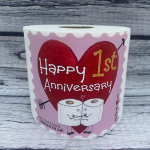 May include: A pink and white toilet paper roll with a red heart and the text "Happy 1st Anniversary" in gold lettering.  The heart has a gold arrow pointing to the "1st".  Two cartoon toilet paper rolls are holding hands below the heart.  The text "One year down, forever to go!" is below the cartoon toilet paper rolls.