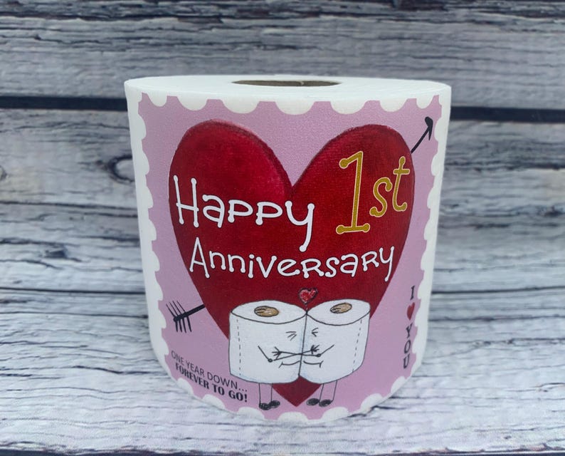 May include: A pink and white toilet paper roll with a red heart and the text "Happy 1st Anniversary" in gold lettering.  The heart has a gold arrow pointing to the "1st".  Two cartoon toilet paper rolls are holding hands below the heart.  The text "One year down, forever to go!" is below the cartoon toilet paper rolls.