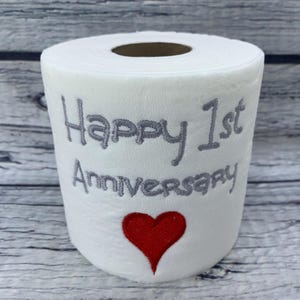 May include: White toilet paper roll with gray embroidered text reading "Happy 1st Anniversary" and a red heart. The roll is on a wooden surface.