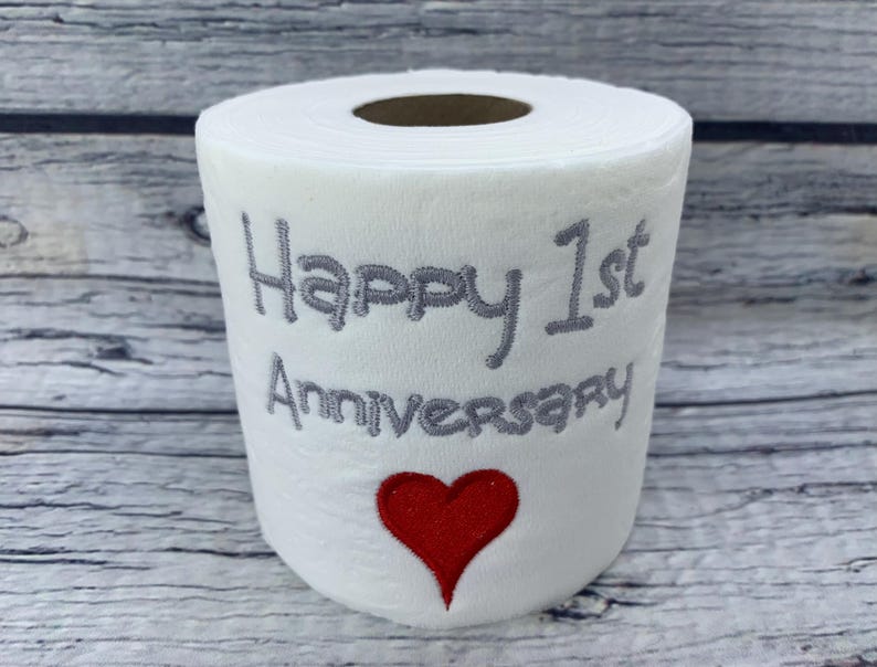 May include: White toilet paper roll with gray embroidered text reading "Happy 1st Anniversary" and a red heart. The roll is on a wooden surface.