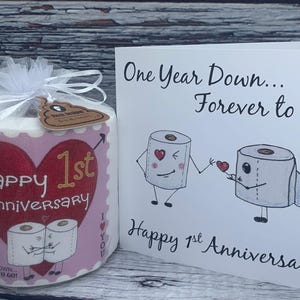 May include: A pink and white toilet paper roll with a red glitter heart and the text "Happy 1st Anniversary" and "One Year Down Forever to Go!"  A card with the text "One Year Down... Forever to go!" and "Happy 1st Anniversary" with a cartoon illustration of two toilet paper rolls holding hands.