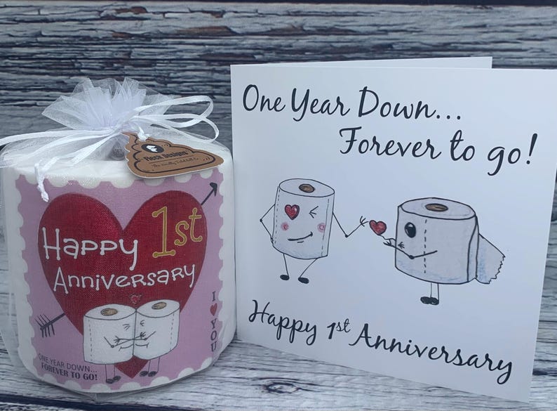 May include: A pink and white toilet paper roll with a red glitter heart and the text "Happy 1st Anniversary" and "One Year Down Forever to Go!"  A card with the text "One Year Down... Forever to go!" and "Happy 1st Anniversary" with a cartoon illustration of two toilet paper rolls holding hands.