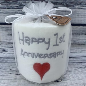 May include: A white, cylindrical gift wrapped in sheer white fabric and tied with a bow. The gift is embroidered with the words "Happy 1st Anniversary" in gray, and a red heart. A small tag is attached.