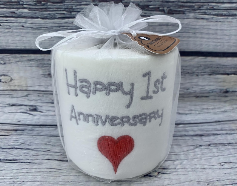 May include: A white, cylindrical gift wrapped in sheer white fabric and tied with a bow. The gift is embroidered with the words "Happy 1st Anniversary" in gray, and a red heart. A small tag is attached.