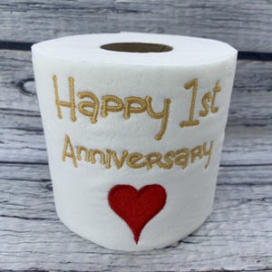 May include: White toilet paper roll with gold embroidered text reading "Happy 1st Anniversary" and a red heart. The roll is a novelty item, likely a gift for a first wedding anniversary.