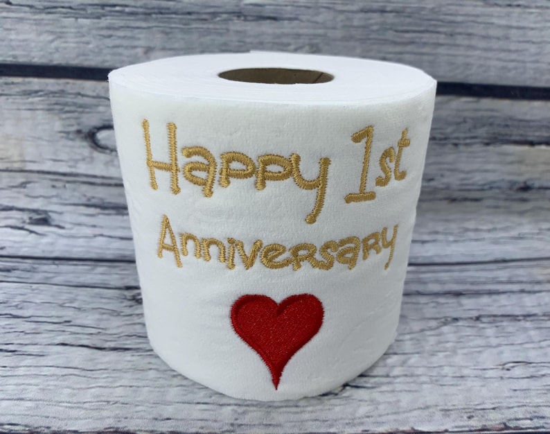 May include: White toilet paper roll with gold embroidered text reading "Happy 1st Anniversary" and a red heart. The roll is a novelty item, likely a gift for a first wedding anniversary.