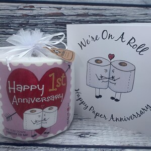 May include: A pink and white toilet paper roll with a heart and the text "Happy 1st Anniversary" and "One year down...forever to go!"  A white card with the text "We're on a roll" and "Happy Paper Anniversary" with a cartoon image of two toilet paper rolls holding hands.