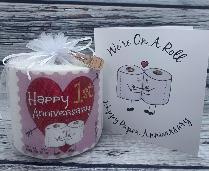 May include: A pink and white toilet paper roll with a heart and the text "Happy 1st Anniversary" and "One year down...forever to go!"  A white card with the text "We're on a roll" and "Happy Paper Anniversary" with a cartoon image of two toilet paper rolls holding hands.